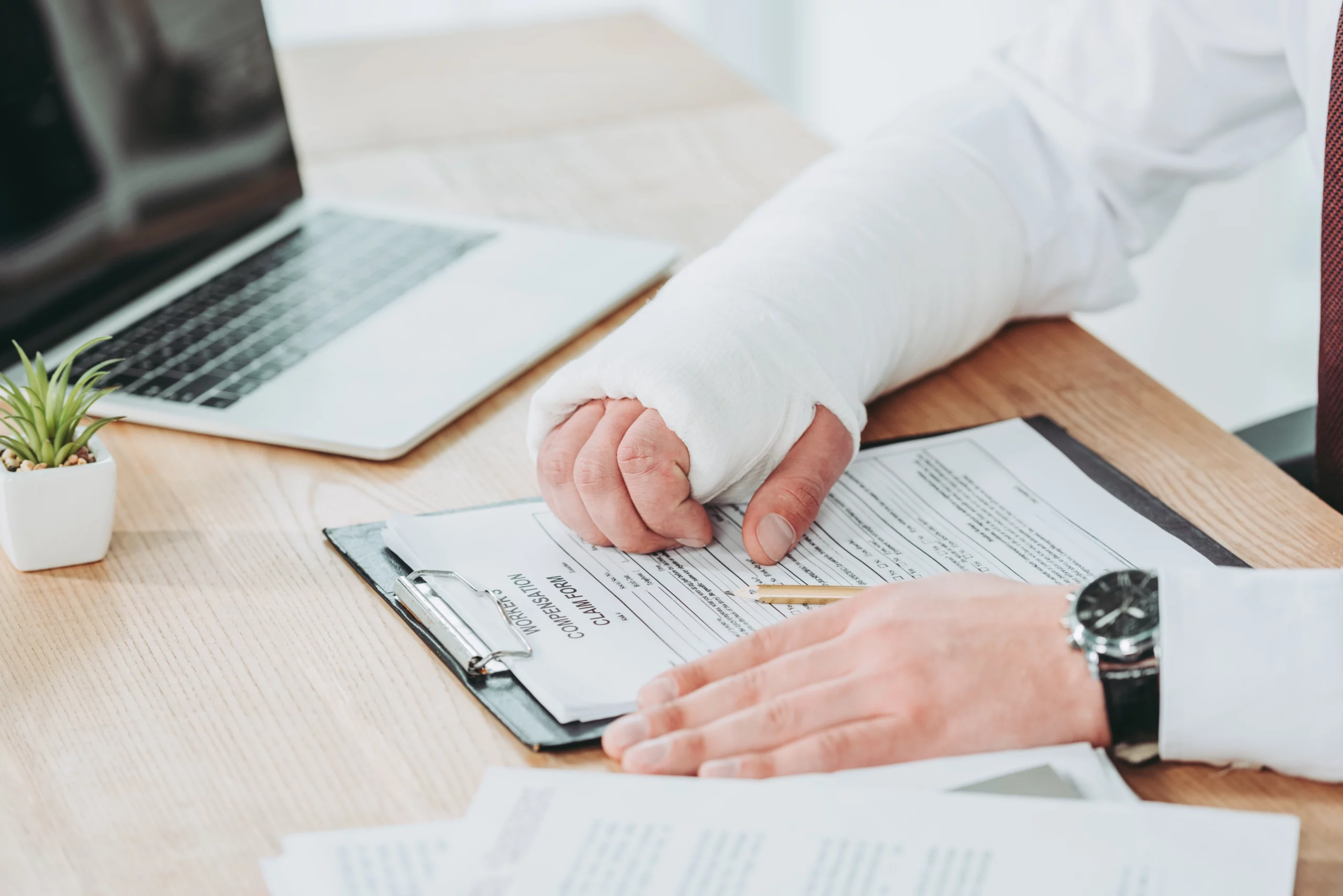 Person with a bandaged arm signing a compensation claim form on a wooden desk beside a laptop and small plant.